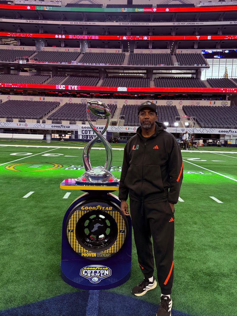 Mike Rumph in a black Miami Hurricanes track suit and cap stands on the field at AT&T Stadium beside the Goodyear Cotton Bowl Classic trophy